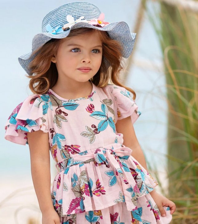 a little girl wearing a floral Mia Belle dress and hat on the beach